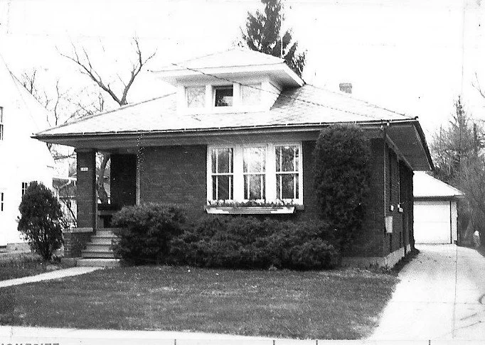 Black-and-white photo of a brick single-story bungalow with a front porch, dormer window, shrubs across the front lawn, and a driveway leading to a detached garage.