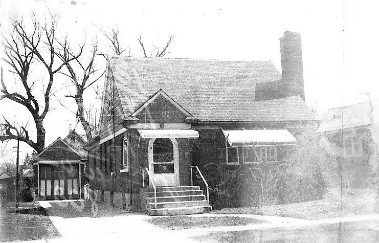 Black-and-white photo of a one-story brick bungalow with front steps and metal awnings, a detached garage to the left and large leafless trees behind.