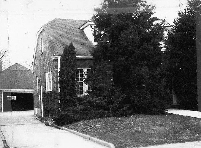 Black-and-white photo of a two-story suburban house partly hidden by tall evergreen trees, with a driveway leading to a garage and a small front lawn.