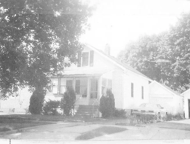 Faded black-and-white photo of a small wood-frame house with a front porch, driveway and parked car, shaded by large trees.