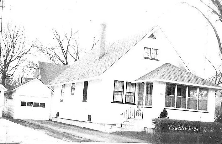 Black-and-white photo of a single-story suburban bungalow with a screened front porch and detached garage.