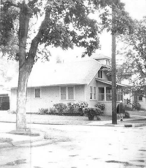 Black-and-white photo of a small bungalow on a tree-lined street corner with two people walking on the sidewalk.