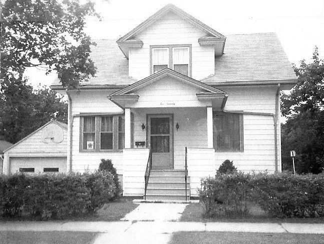 Black-and-white photo of a two-story wooden house with a front porch and steps, flanked by trimmed shrubs and a detached garage to the left.