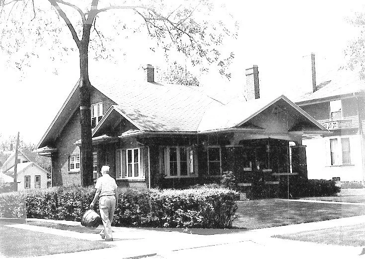 Black-and-white photo of a single-story bungalow with a front porch; a man walks on the sidewalk carrying a lawn mower bag.