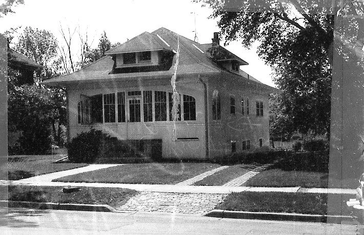 Black-and-white photo of a two-story bungalow with an enclosed front porch, driveway, sidewalk, and surrounding trees.