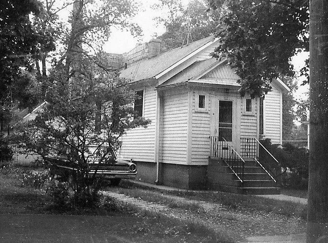 Black-and-white photo of a small wooden bungalow with front steps, surrounded by trees and a partially visible parked car.