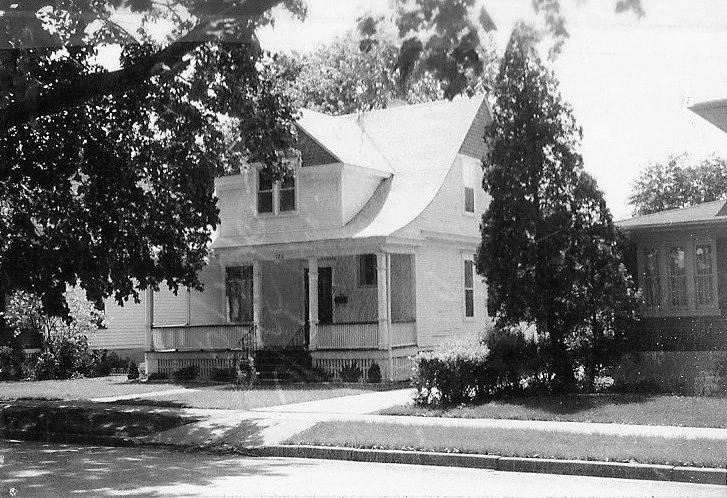 Black-and-white photo of a two-story house with a covered front porch, large trees and a sidewalk in the foreground.