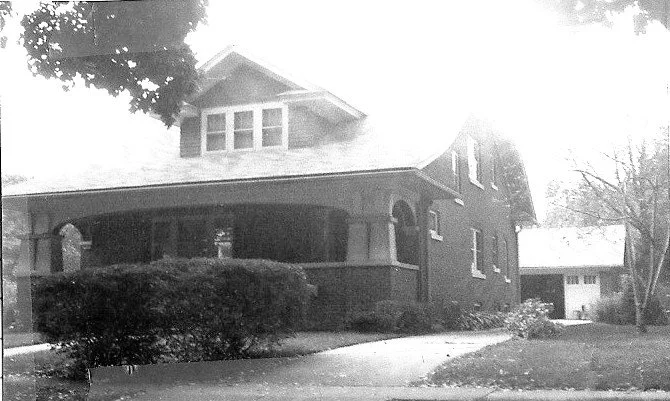 Black-and-white photo of a two-story brick house with a covered front porch, shrubs, driveway, and a detached garage in the background.