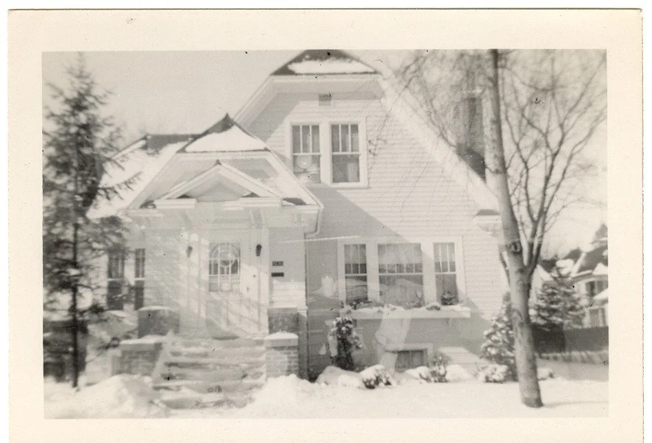 Black-and-white vintage photo of a two-story snow-covered house with a front porch, bare trees, and steps leading up through the snow.