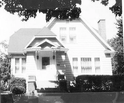 A black-and-white photo of a two-story house with a small front porch, steps, shrubs, and a chimney, framed by tree branches.