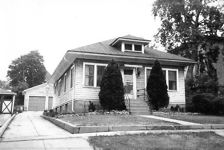 Black-and-white photo of a small single-story bungalow with front steps flanked by two tall shrubs, a driveway leading to a detached garage, and a lawn in front.