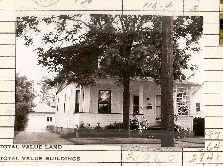Black-and-white photo of a small two-story wood house with a front porch and two large trees, driveway to a detached garage at left, mounted on a valuation form.