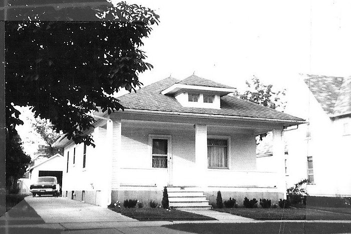 Black-and-white photo of a single-story bungalow with a front porch, steps, small shrubs, and a driveway leading to a garage with a parked car.