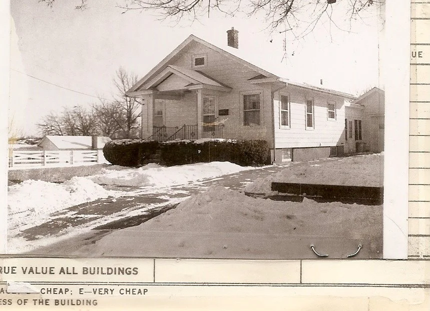 Sepia photo of a single‑story bungalow with front porch and chimney, snowy yard and driveway, trimmed hedge and white fence; paper border visible at bottom.
