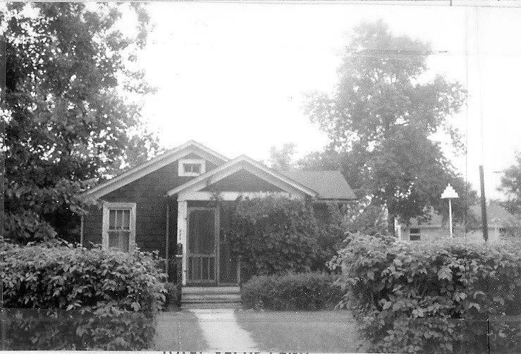 Black-and-white photo of a small single-story house with a screened front porch, overgrown shrubs lining a walkway, and large trees in the background.