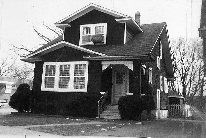 Black-and-white photo of a two-story Craftsman-style house with a front porch, dormer window and small shrubs, viewed from the street. (Copy)