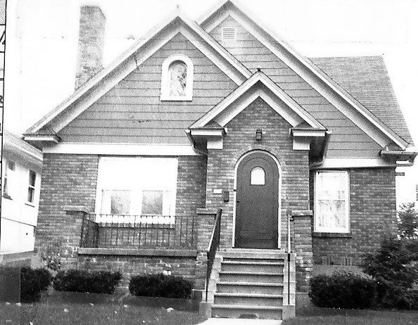 Front view of a small brick bungalow with a central arched door, front steps, and two windows.