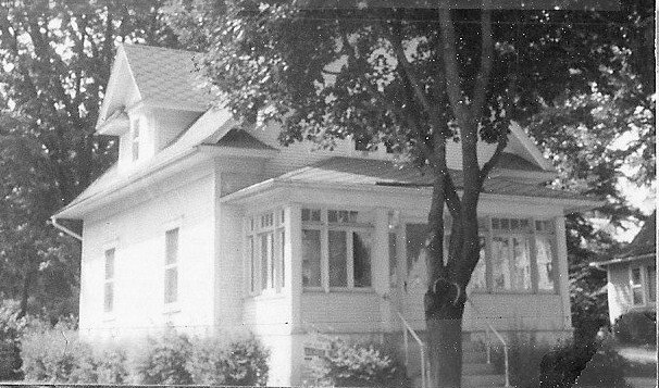 Black-and-white photo of a small two-story house with an enclosed front porch and a tree in front.