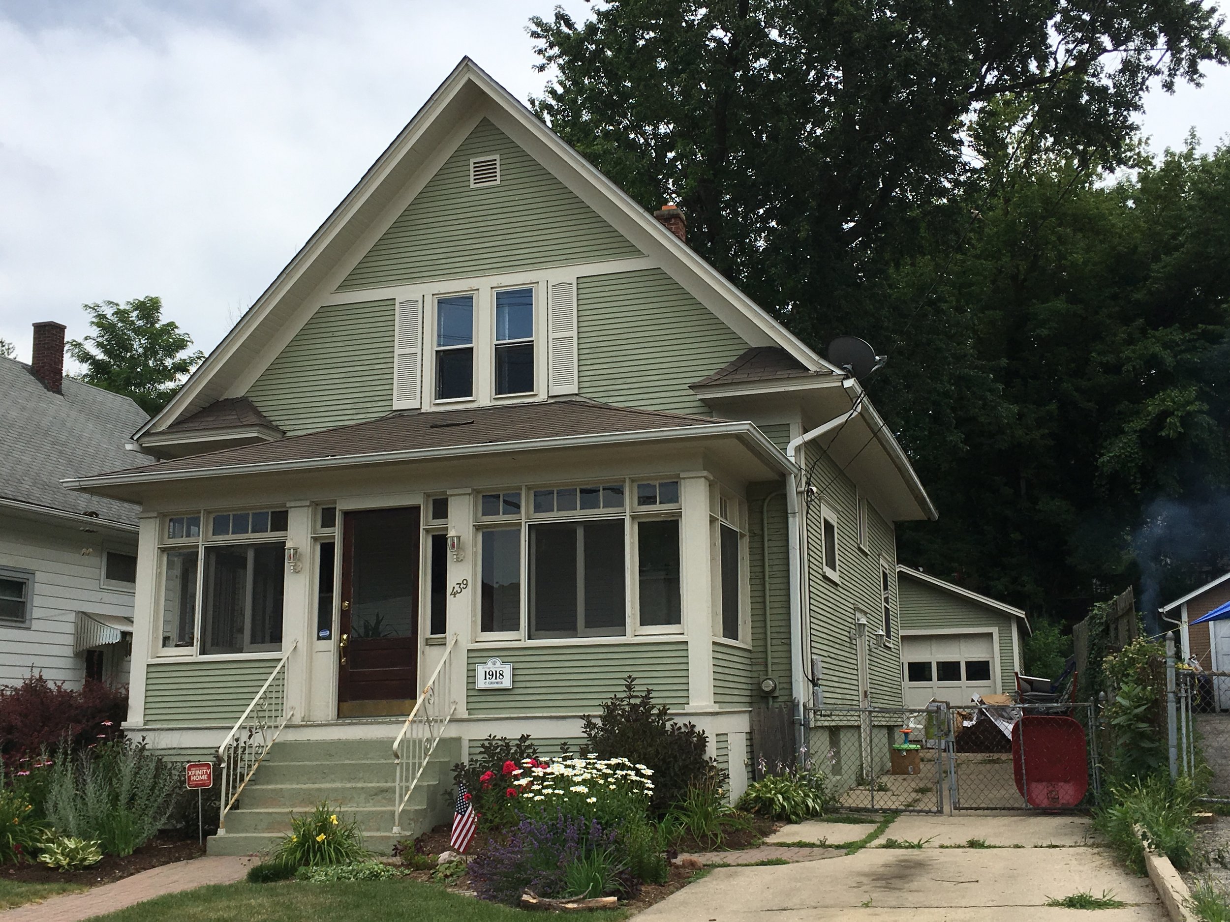 Front view of a green two-story Craftsman-style house with a steep gable, enclosed front porch, flower garden and a small American flag.