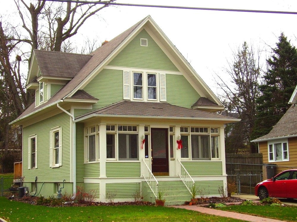 Two-story light green house with an enclosed front porch decorated with red bows and a small lawn; a red car is parked to the right.
