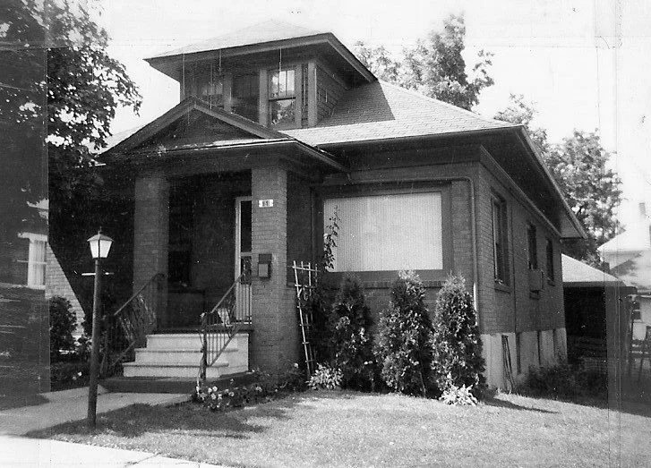Black-and-white photo of a one-story brick bungalow with a covered front porch, steps and railing, lamp post, and shrubs.