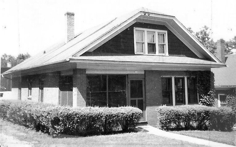 Black-and-white photo of a single-story brick bungalow with a central front door, dormer window, chimney, and trimmed shrubs along the front.