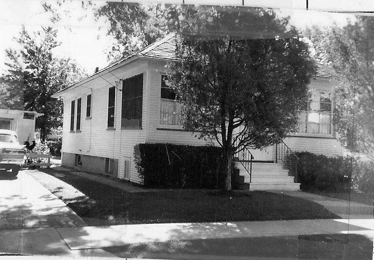 Black-and-white photo of a single-story raised house with front steps, a tree and shrubs in the yard, and a car parked in the driveway.