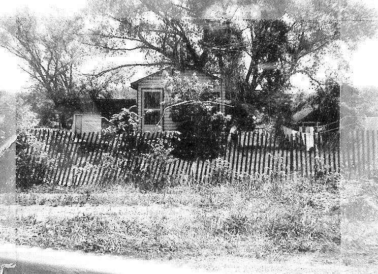 Black-and-white photo of an old house partly hidden by a large tree and a leaning picket fence with overgrown grass.