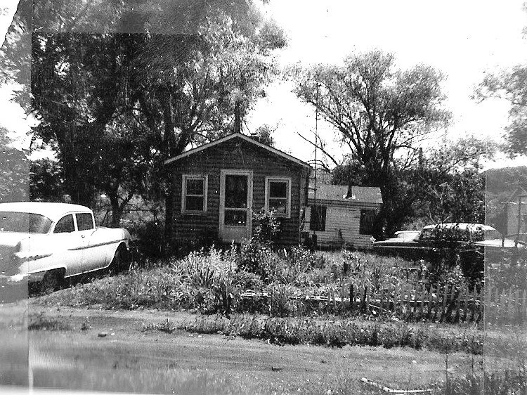 Black-and-white photo of a small wooden bungalow with an overgrown front yard and trees, flanked by two vintage cars parked on either side.