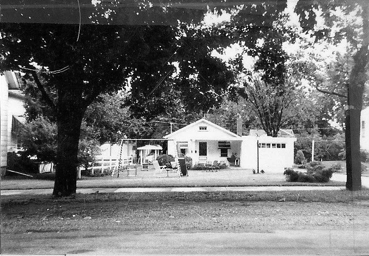 Black-and-white photo of a small single-story house with a front lawn, patio chairs and umbrella, a swing set, and large trees framing the scene.