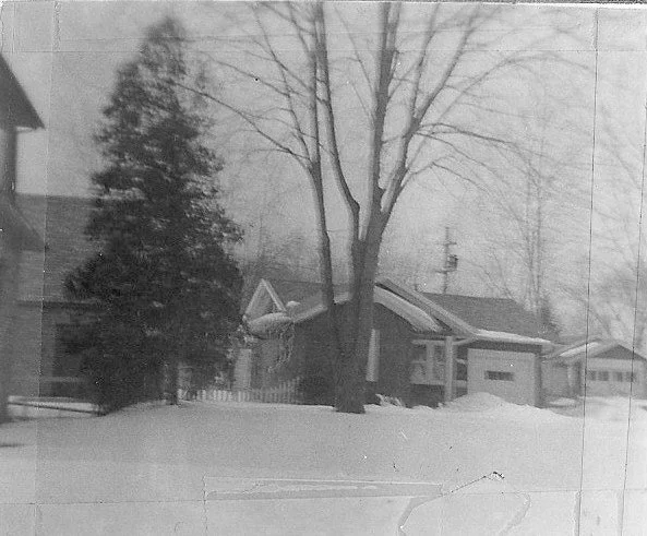 Black-and-white photo of a small house and garage behind a snow-covered yard, with bare trees and an evergreen in the foreground.