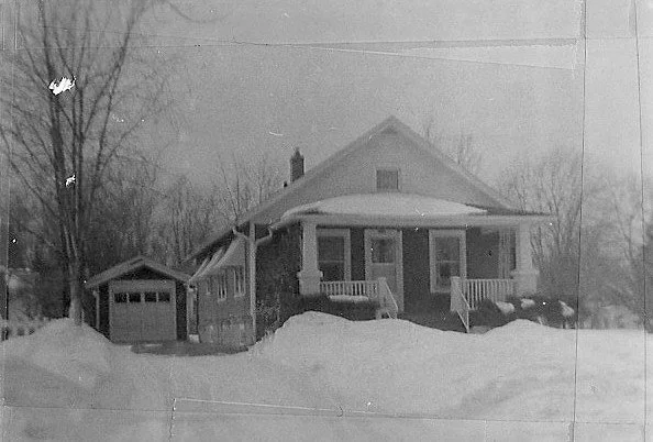 Black-and-white photo of a snow-covered one-story bungalow with a front porch, detached garage to the left, and bare trees in the background.
