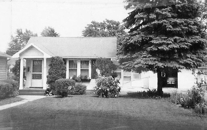 Black-and-white photo of a small single-story house with a manicured lawn, large evergreen tree, and a driveway leading to a detached garage.