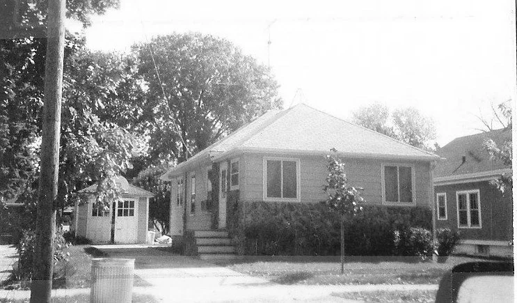 Black-and-white photo of a single-story bungalow with front steps, small yard and detached shed, trees and a street in front.