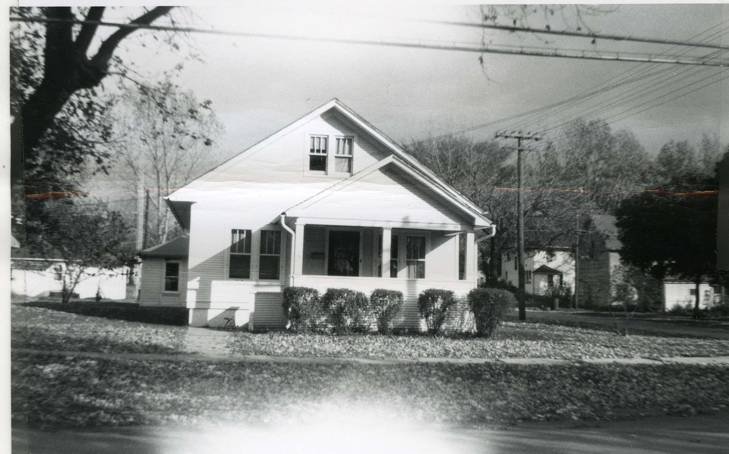 Black-and-white photo of a two-story gabled house with a front porch, trimmed shrubs, and trees in a residential neighborhood.