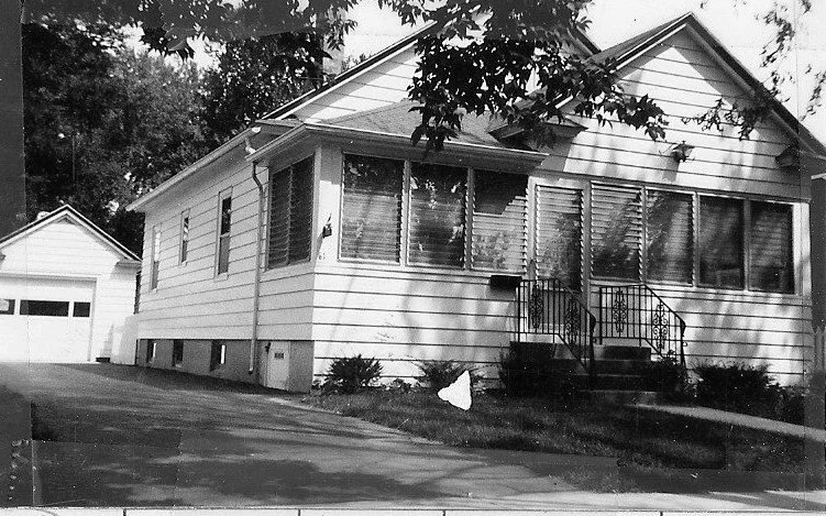 Black-and-white photo of a single-story white siding house with an enclosed front porch, steps with wrought-iron railings, a driveway leading to a detached garage, and tree branches overhead.