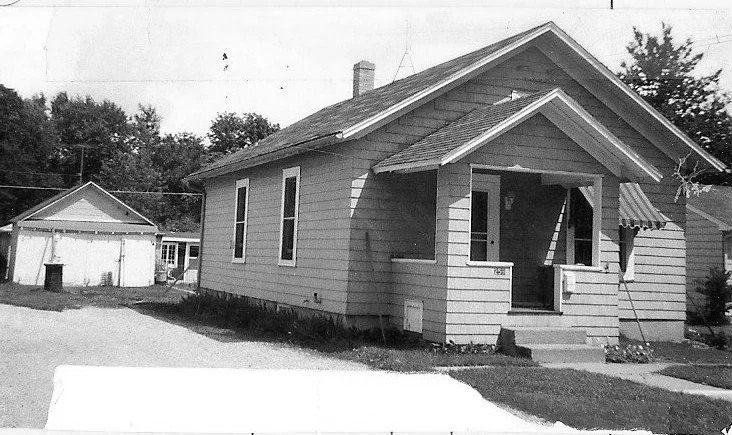Black-and-white photo of a small single-story bungalow with a covered front porch and a detached garage in the backyard.