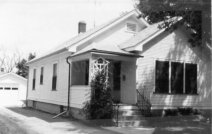 Black-and-white photo of a single-story wood-sided house with a small covered front porch, concrete steps and a driveway leading to a detached garage.