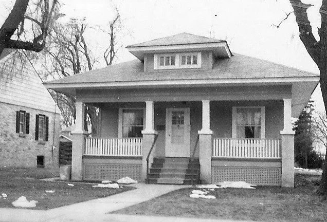 Black-and-white photo of a one-story bungalow with a covered front porch, central doorway and steps, flanked by windows and small patches of snow on the lawn.