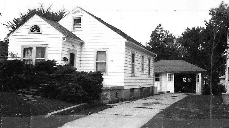 Alt text: "Small white single-story house with siding, front shrubs and a driveway leading to a detached garage."