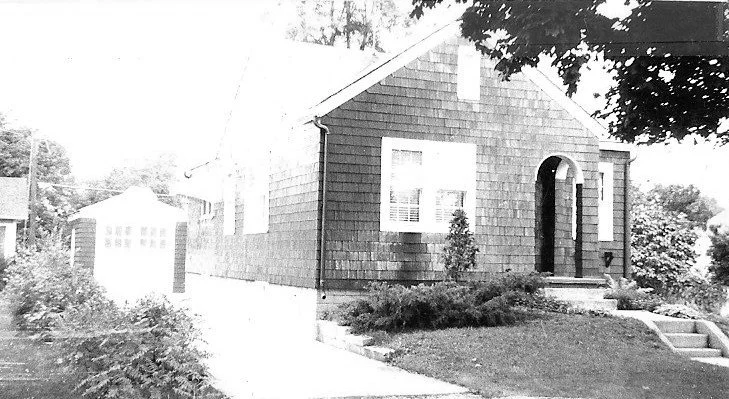 Small shingled bungalow with an arched front doorway, front lawn and walkway, and a driveway leading to a detached garage.
