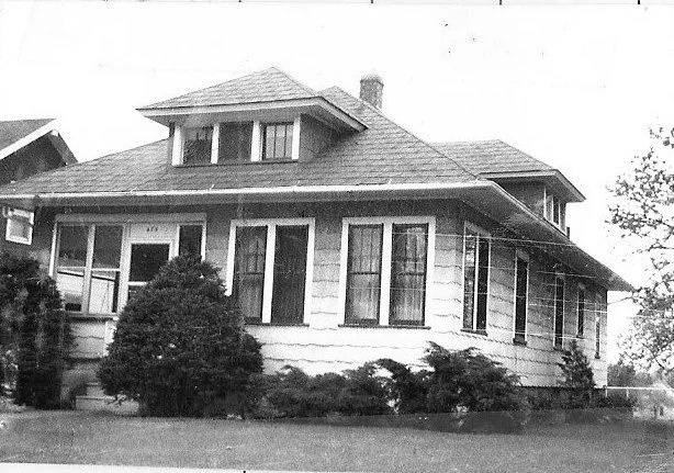 Black-and-white photo of a single-story bungalow-style house with a front porch, dormer window, chimney and shrubs in the yard.
