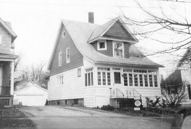 Black-and-white photo of a two-story house with an enclosed front porch, steep gabled roof, driveway, and detached garage.