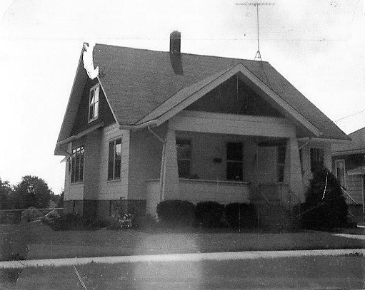 Black-and-white photo of a single-story bungalow with a steep gabled roof, front porch, and shrubs along the foundation.