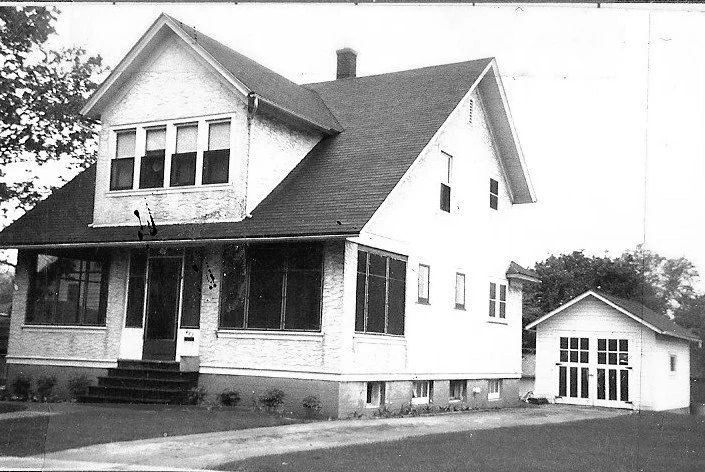 Black-and-white photo of a two-story house with an enclosed front porch and a detached garage to the right.