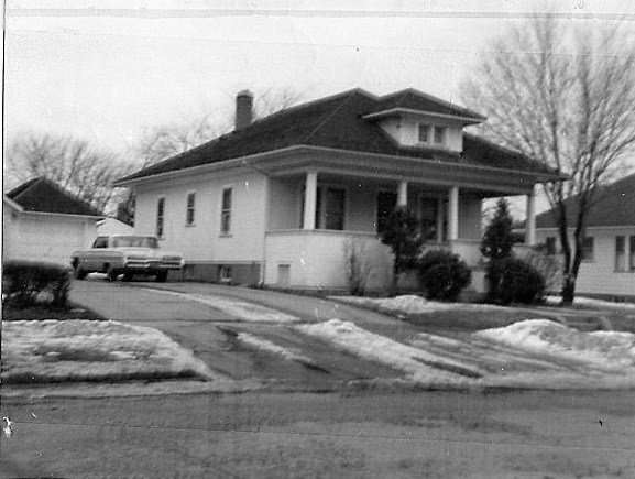 Black-and-white photo of a single-story bungalow with a covered front porch, a vintage car parked in the driveway, leafless trees, and patches of snow on the lawn.
