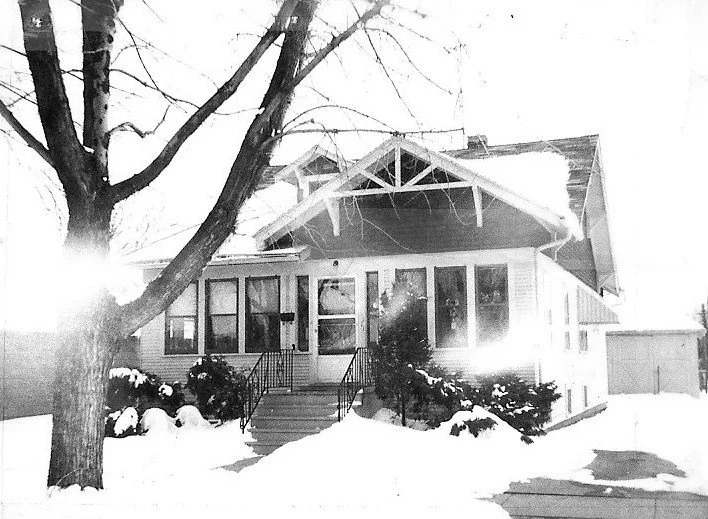 Black-and-white photo of a snow-covered bungalow with a front enclosed porch, steps, shrubs, and a bare tree in the yard.