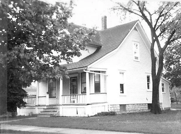 Black-and-white photo of a two-story wood-frame house with a covered front porch, white railing, and trees on either side.