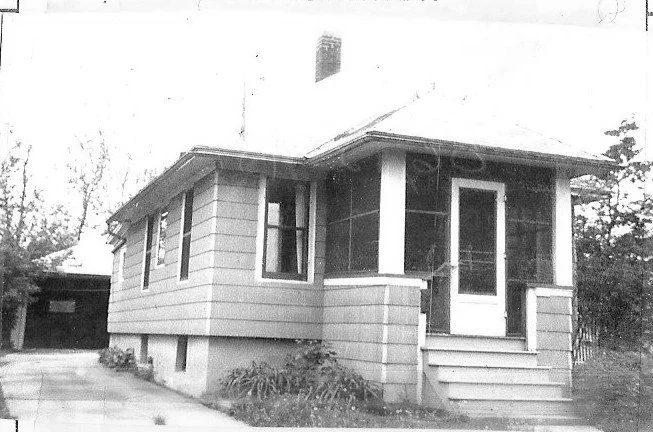 Black-and-white photo of a small single-story bungalow with a screened front porch and steps, driveway to the left leading to a detached garage.