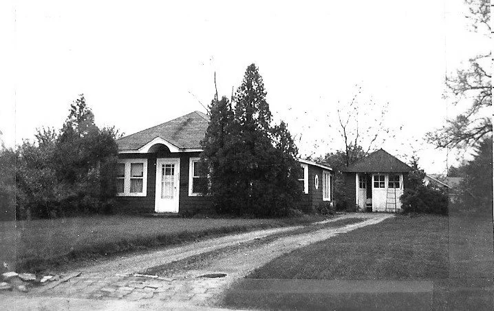 Black-and-white photo of a single-story bungalow with a gravel driveway, trees in front, and a small detached garage to the right.